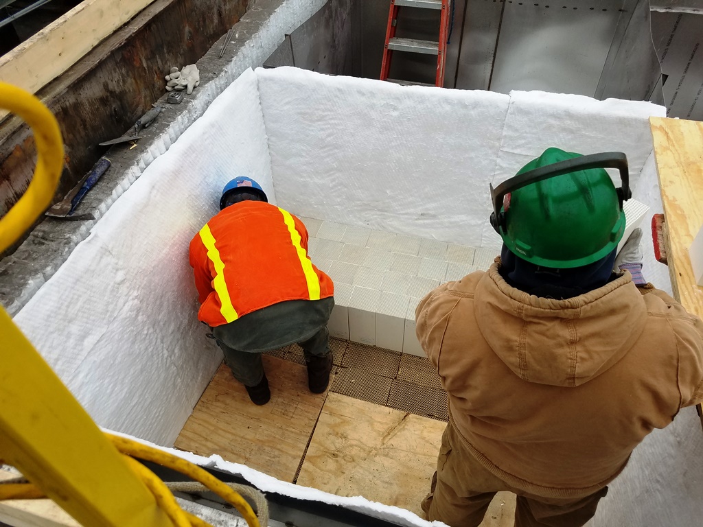 Two construction workers in high-vis gear and jackets work inside an individual can, installing ceramic media blocks and ceramic fiber blanket lining.