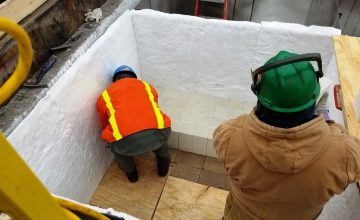 Two construction workers in high-vis gear and jackets work inside an individual can, installing ceramic media blocks and ceramic fiber blanket lining.